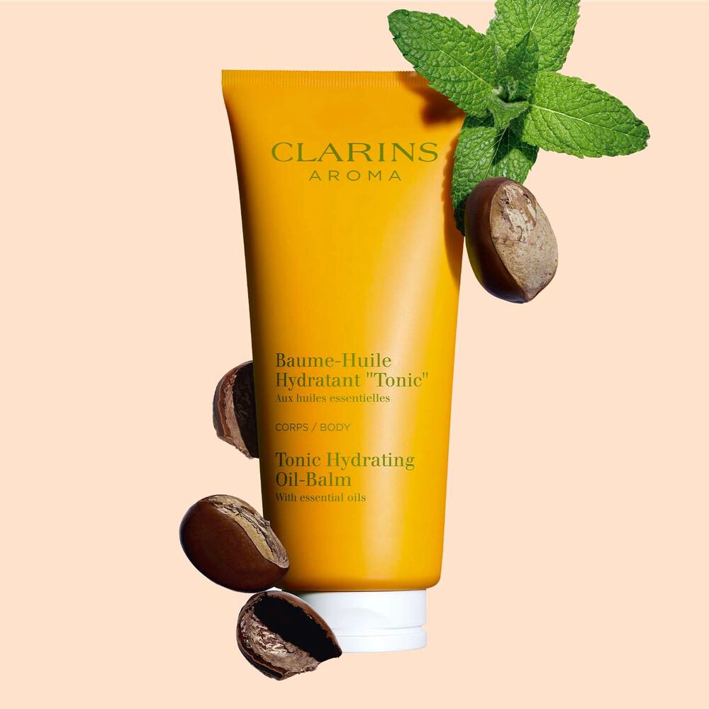 Packshot of a tube of Clarins Tonic Hydrating Oil-balm, close to some mint leaves and shea nuts on a peach background.