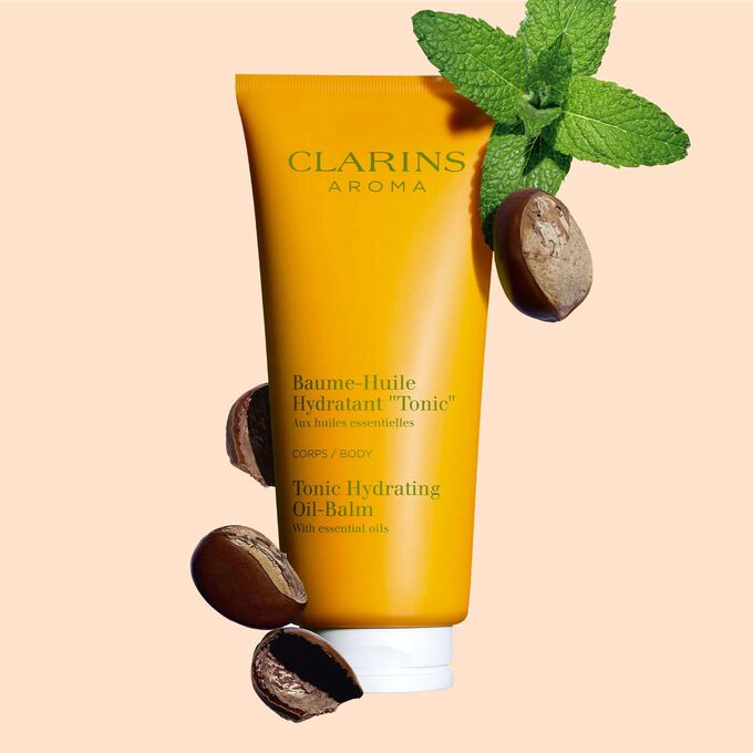 Packshot of a tube of Clarins Tonic Hydrating Oil-balm, close to some mint leaves and shea nuts on a peach background.